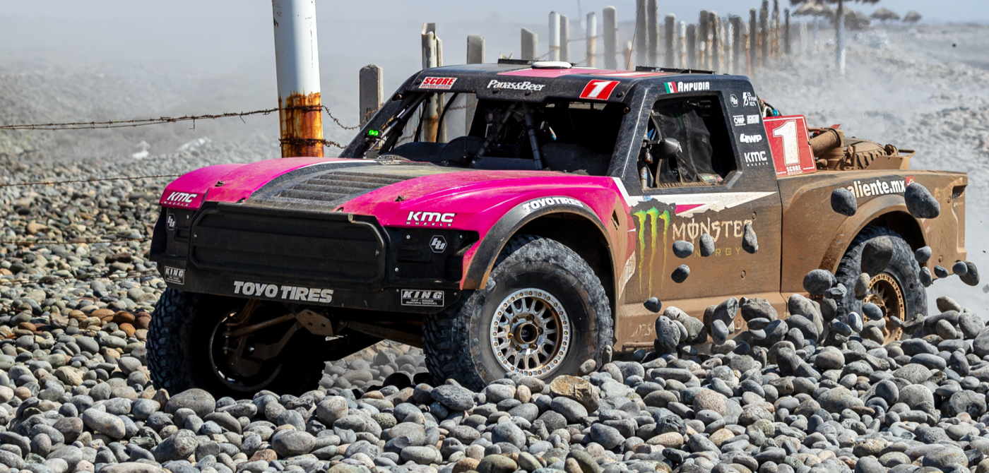 Pink and black off-road vehicle on a rocky beach with promotional text about the 2025 SCORE Baja 500.